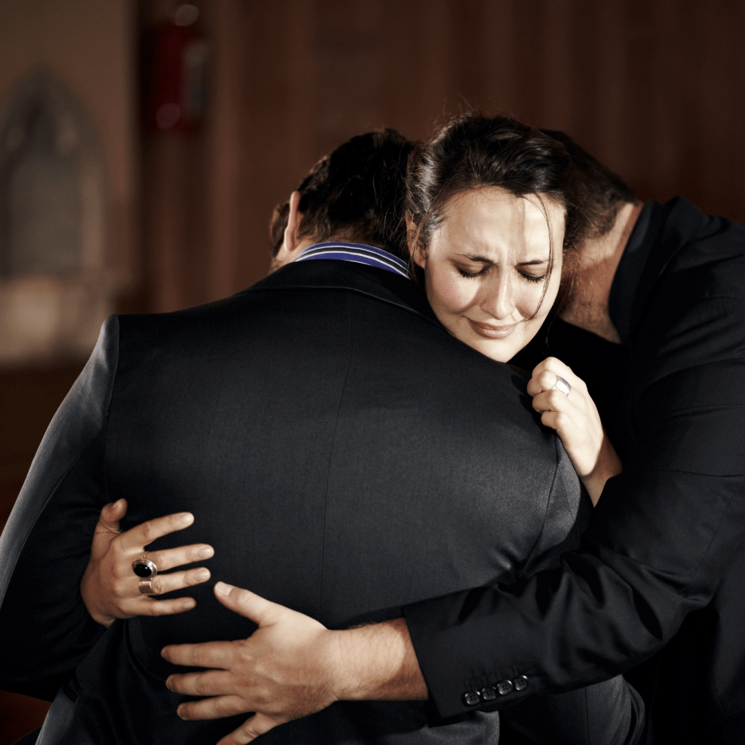 Zwei Herren nehmen eine Dame in den Arm und trösten sie auf einer Beerdigung